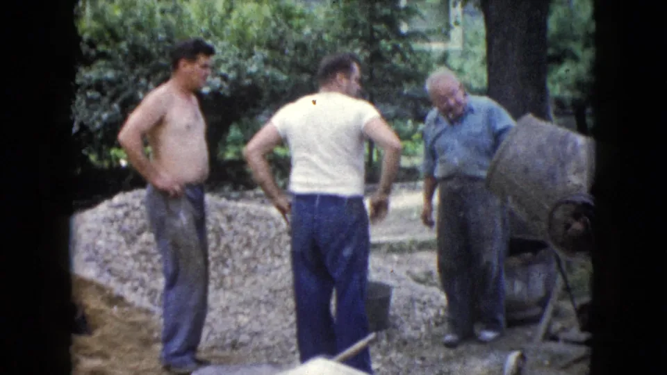 Three adult men are seen beside a cement mixer and gravel pile in 1960 Wauconda, Illinois, with trees and yard equipment nearby.