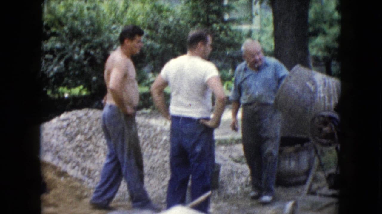 Three men stand beside a concrete mixer and gravel pile in an outdoor yard.