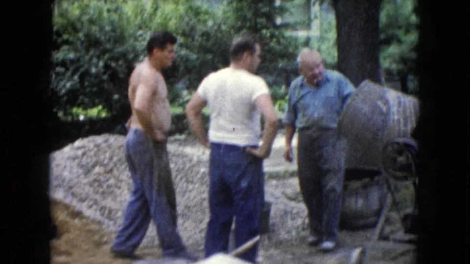In Wauconda, Illinois in 1960, two younger men and an older man stand near a concrete mixer and aggregate pile during outdoor work.