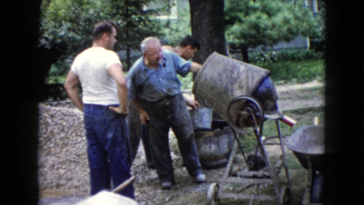 In Wauconda, Illinois, 1960, three adult men gather around a cement mixer near gravel and trees in a backyard.