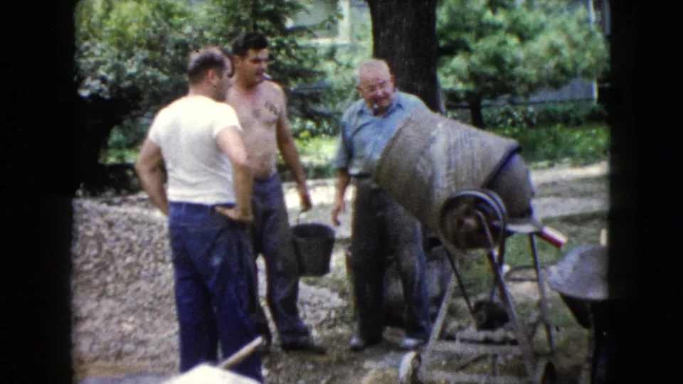 Three adult men in a suburban yard stand around a concrete mixer, one shirtless and another holding or standing near a bucket.