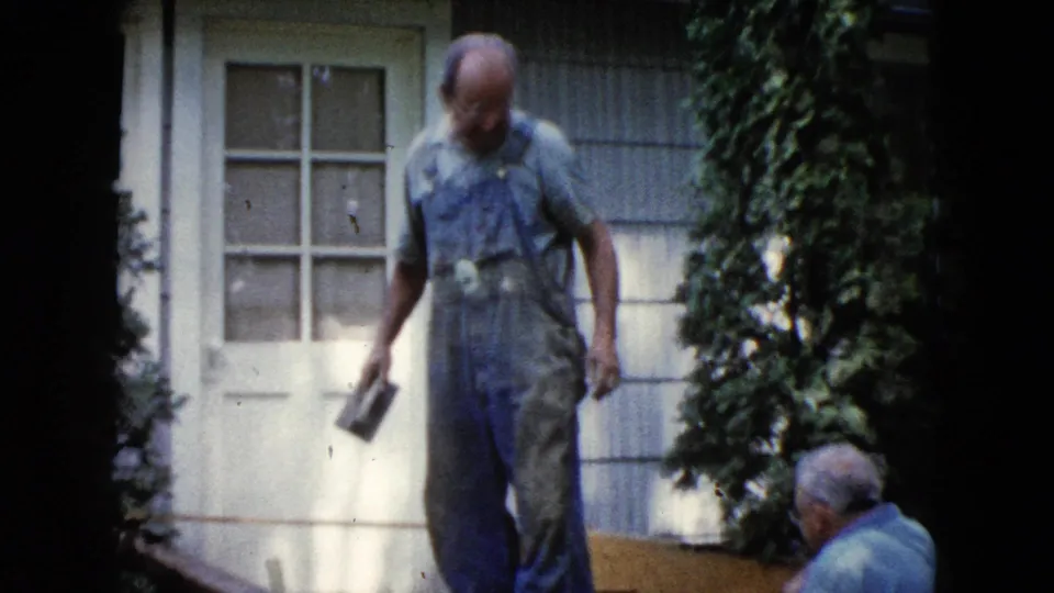 A man in blue overalls walks with a masonry tool near a white door and shrubs in the Wauconda home-movie scene.