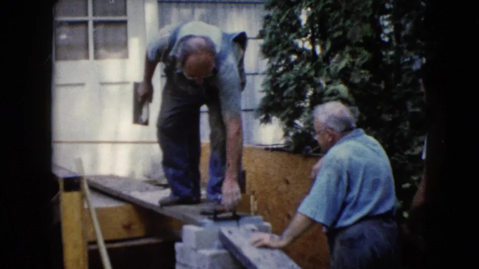 One man bends over a wooden form with a tool while another adult man sits nearby beside cinder blocks in the 1960 Wauconda footage.