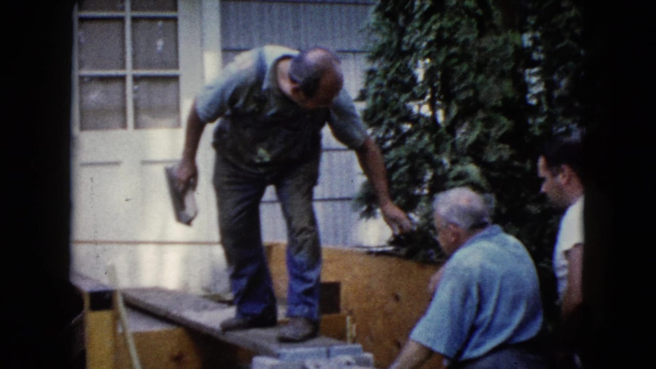 Man in overalls uses a trowel near concrete blocks while two men sit beside a house.
