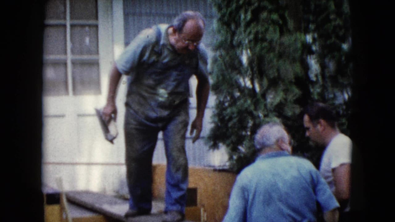A man in blue overalls stands on a plank near a white door, holding a trowel while two men sit beside the outdoor work area.