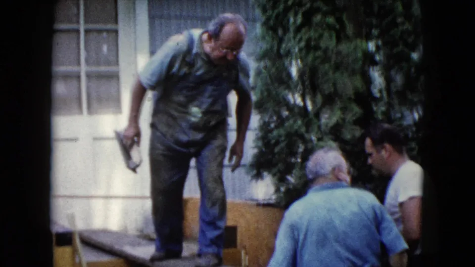 A man in blue overalls stands on a plank near a white door, holding a trowel while two men sit beside the outdoor work area.