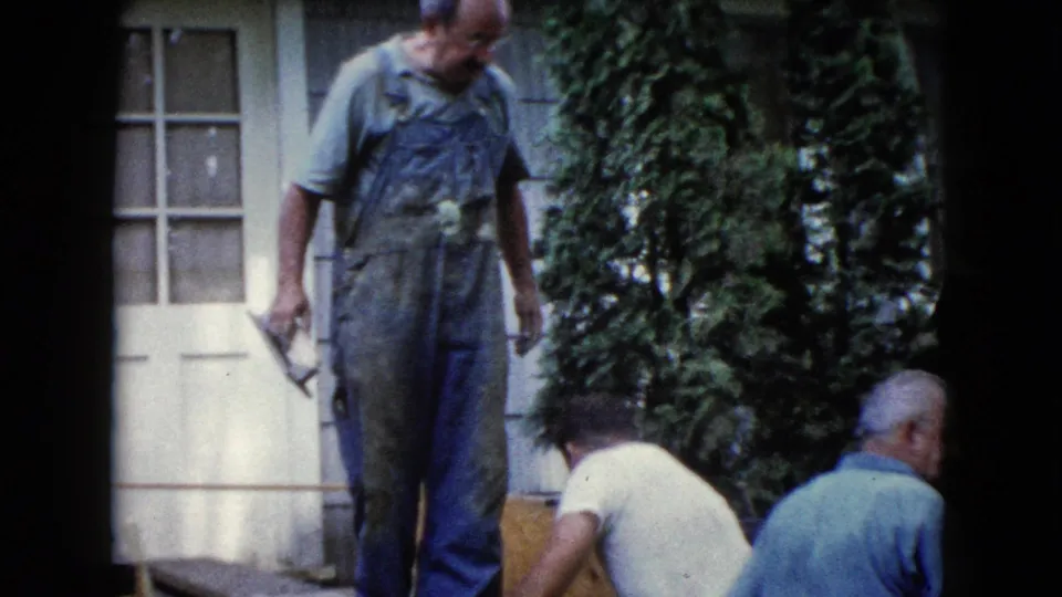 In the Wauconda home-movie scene, a worker in overalls stands with a trowel as two men lean near the work area by a white exterior door.