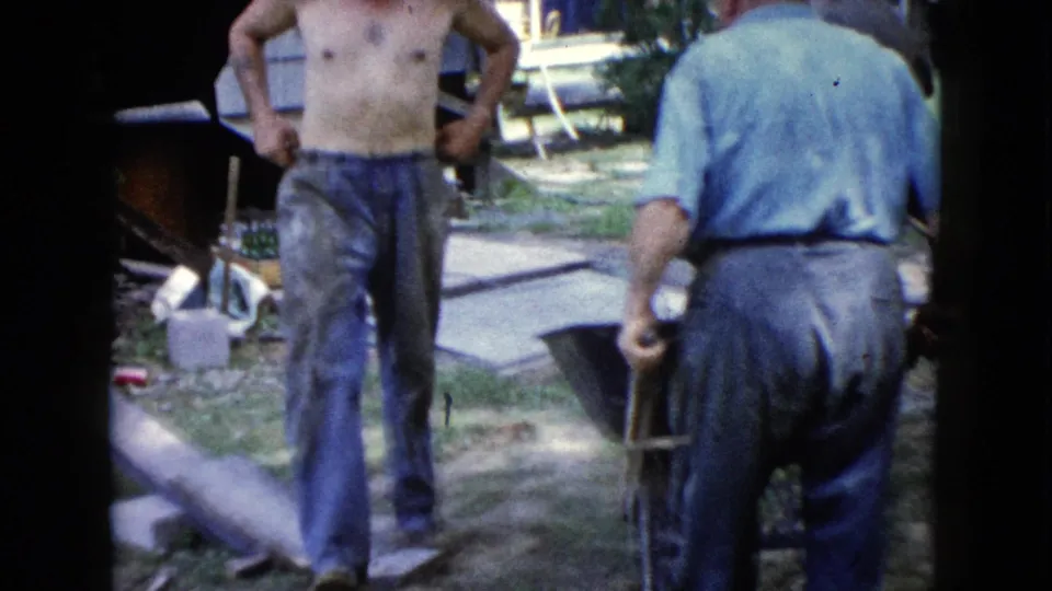 Two men stand near concrete blocks, tools, and a plank in a yard beside a house, continuing the 1960 Wauconda home-work scene.