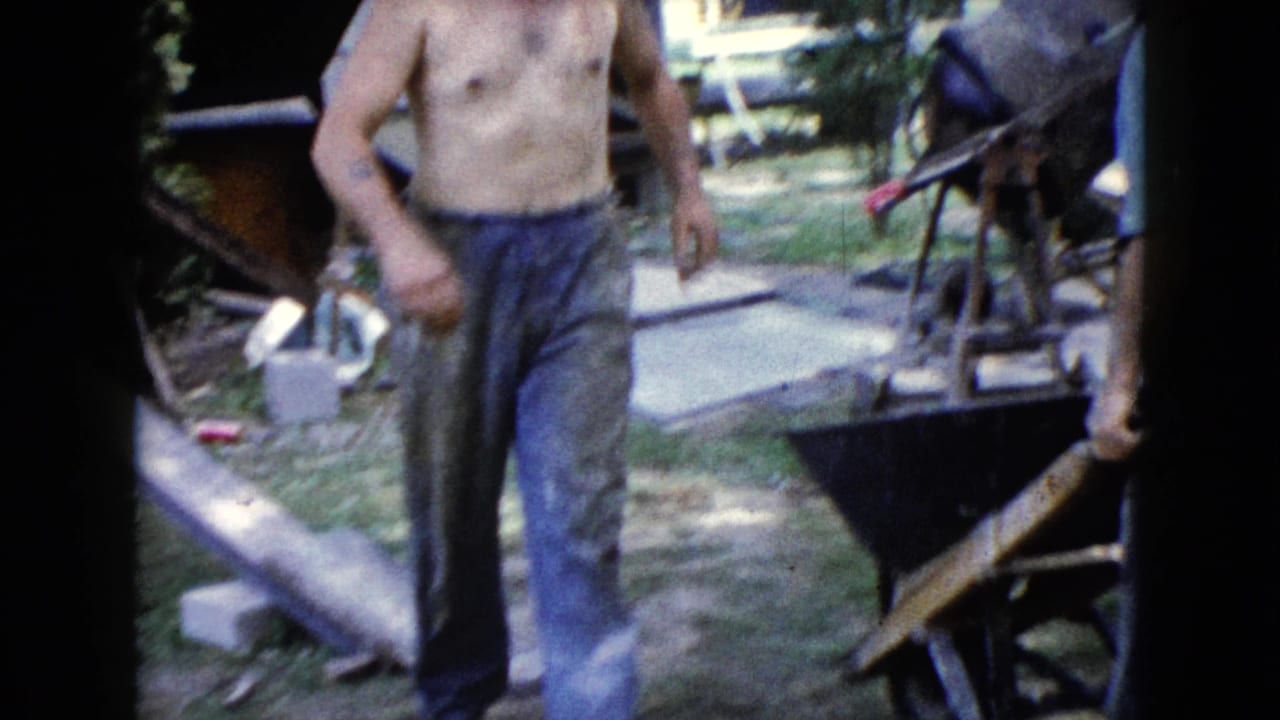 Shirtless man in jeans walks near wheelbarrows and backyard construction materials.