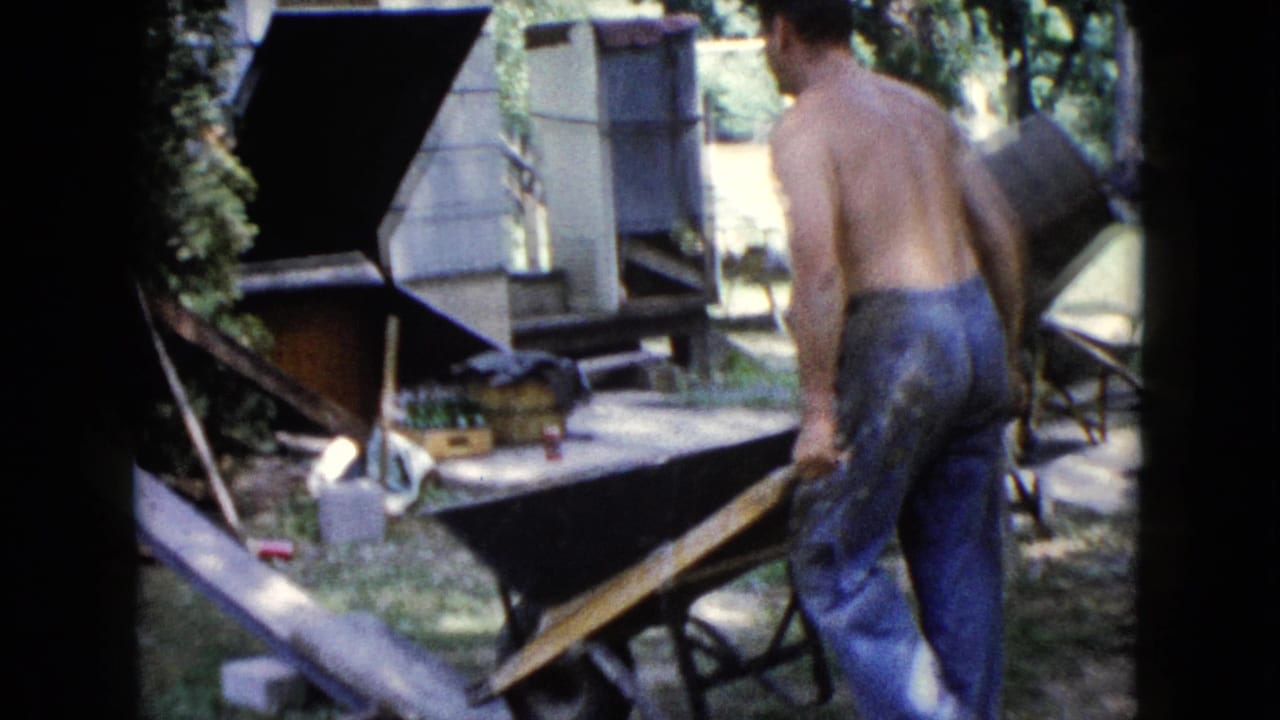1960 Wauconda frame of a shirtless man handling a wheelbarrow near a house wall and yard materials.