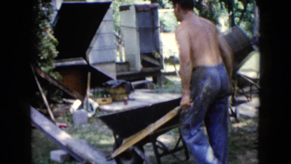 1960 Wauconda frame of a shirtless man handling a wheelbarrow near a house wall and yard materials.