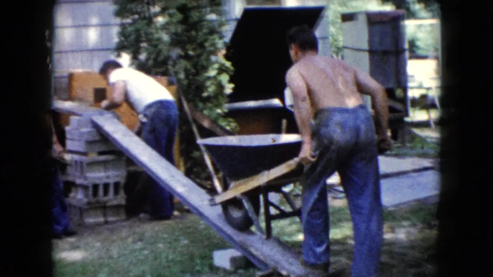 1960 Wauconda backyard construction frame with two men near cinder blocks and a shirtless man moving a wheelbarrow.