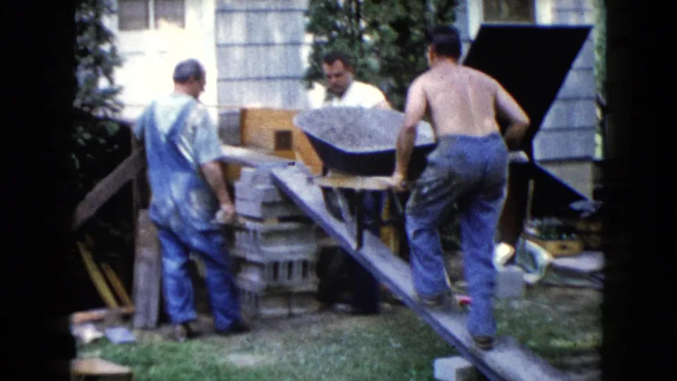 1960 Wauconda home-movie frame showing three adult men working with a loaded wheelbarrow, blocks, and a wooden ramp.