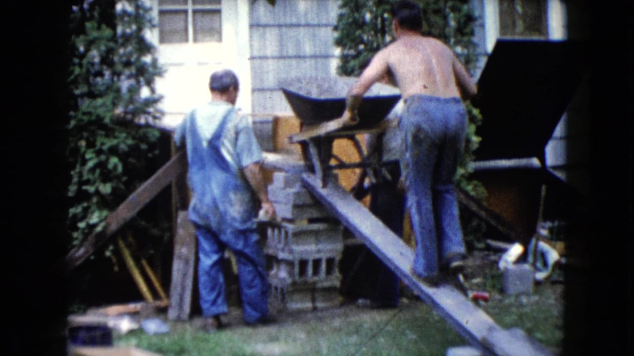 Three men work near cinder blocks as one pushes a wheelbarrow up a plank ramp.