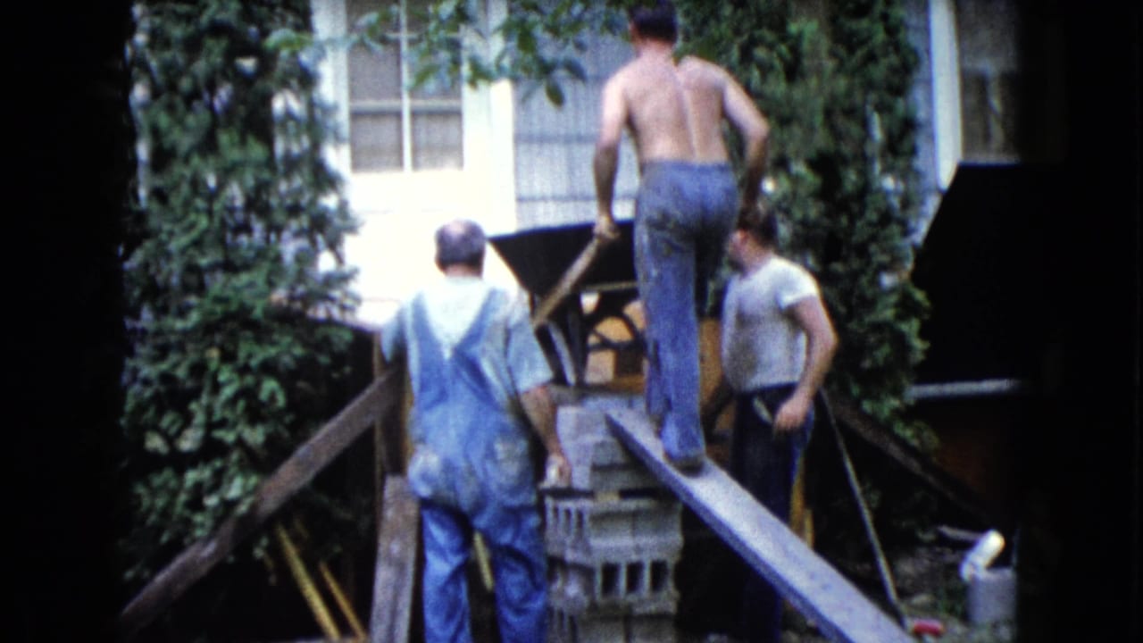 A shirtless man moves a wheelbarrow up a plank while two men stand nearby.