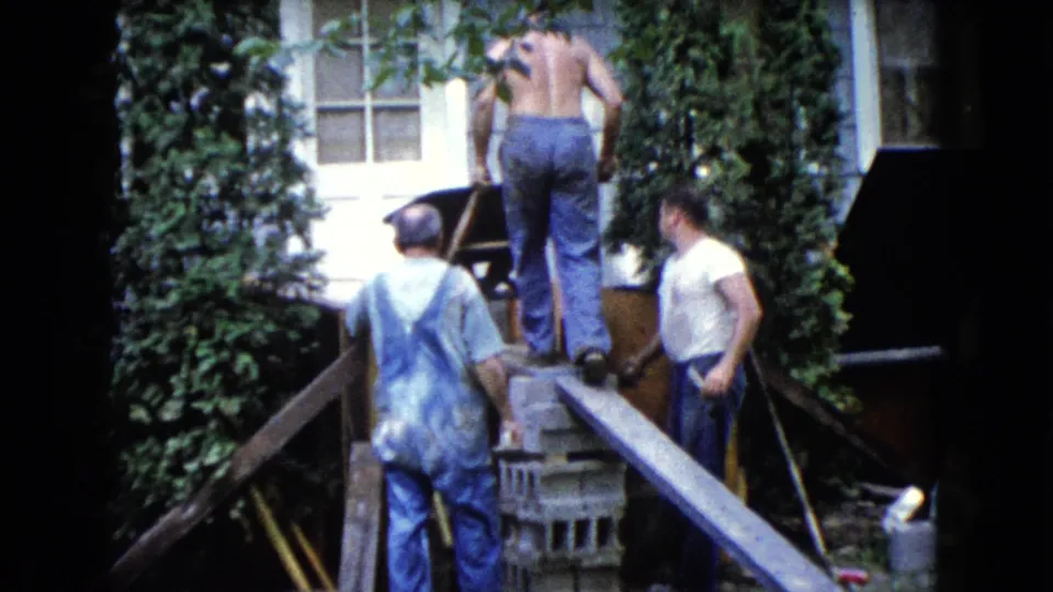 The home-movie frame shows three adult men, stacked cinder blocks, a wheelbarrow, and a plank ramp beside a residential exterior in 1960.
