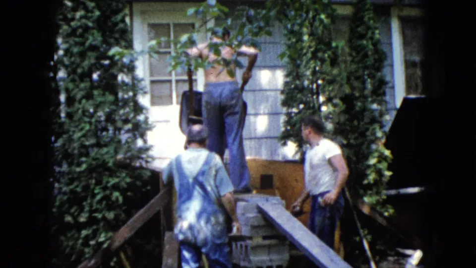 The 8mm frame shows three adult men in a backyard work area beside an ivy-covered Wauconda house exterior.