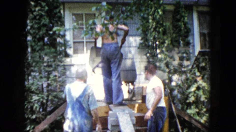 A bright 1960 8mm frame shows three men working at a Wauconda house exterior, partly washed by sunlight and framed by ivy.