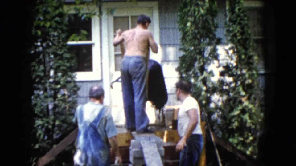1960 Wauconda home-movie frame of men assisting a shirtless worker on a makeshift platform outside an ivy-covered house.