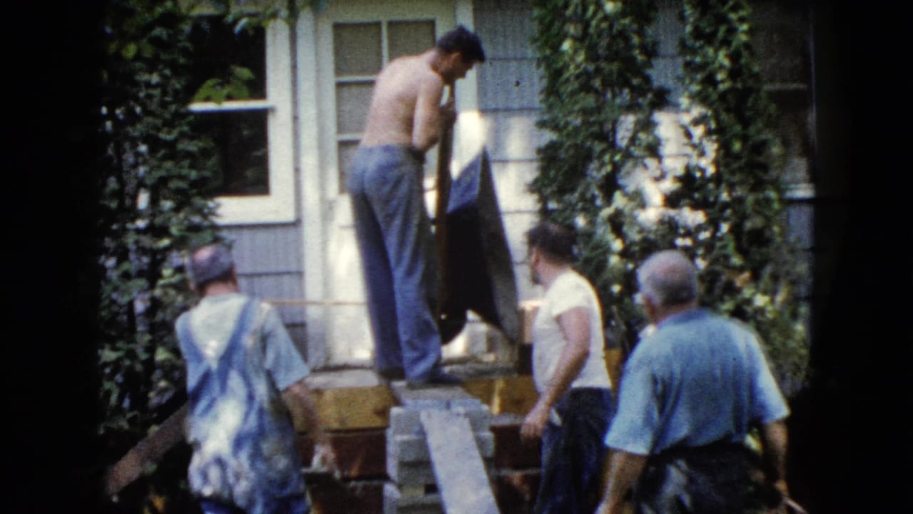 Four men stand near porch steps as a shirtless man uses a shovel by a house door.