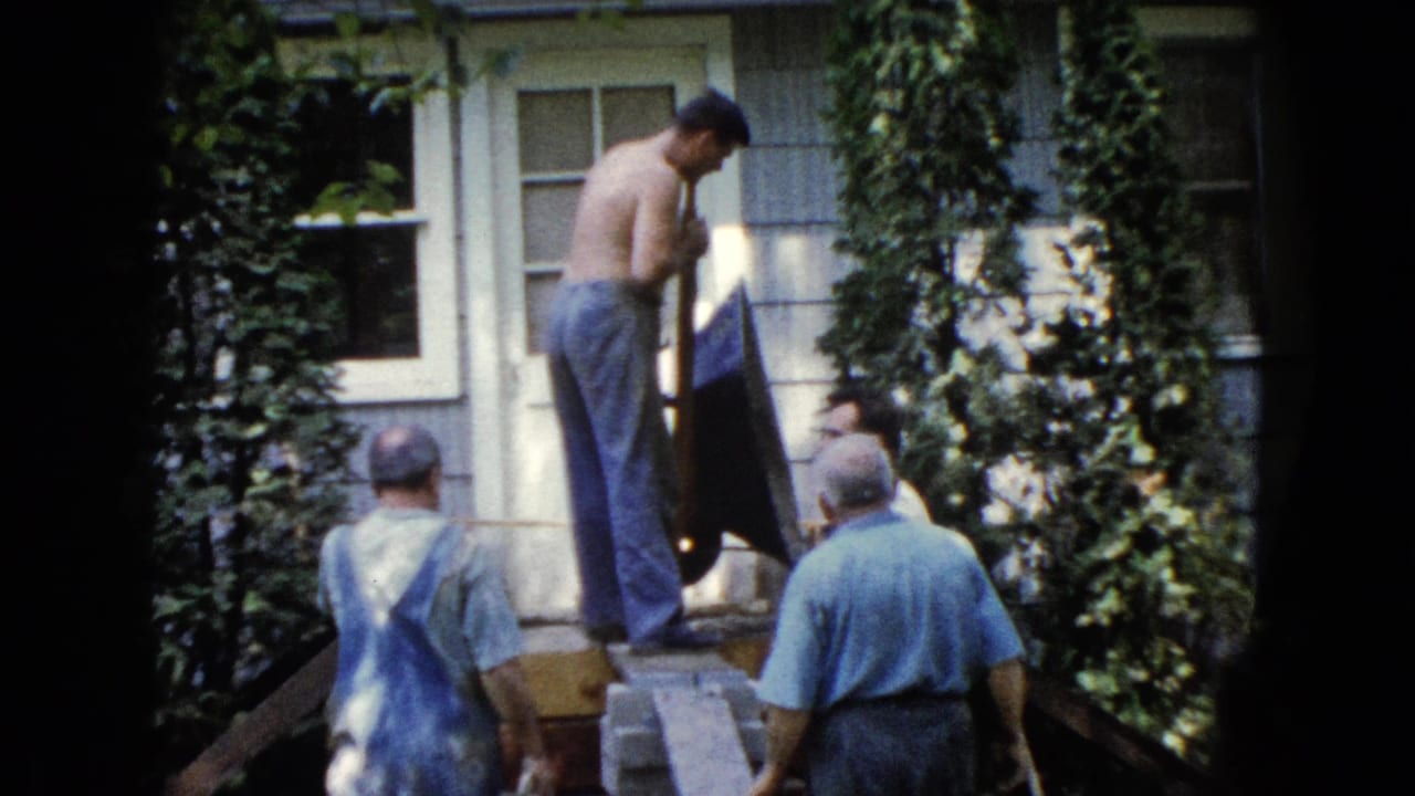 A 1960 Wauconda frame shows men in casual work clothes around porch steps as a shirtless man handles a shovel beside a house door.