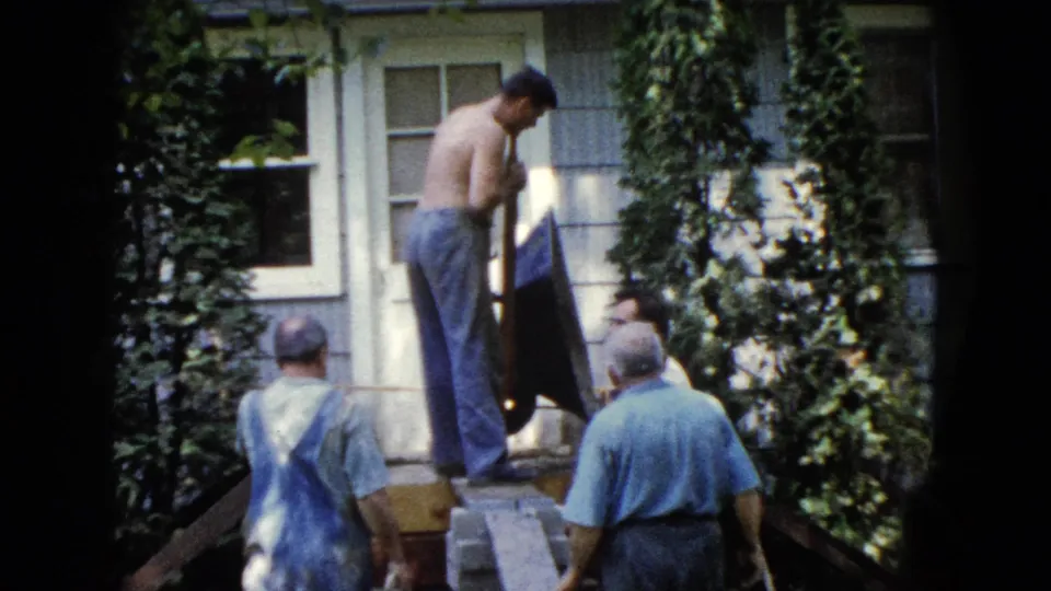 A 1960 Wauconda frame shows men in casual work clothes around porch steps as a shirtless man handles a shovel beside a house door.