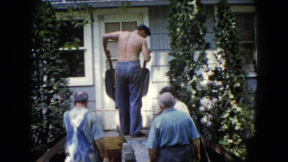 In this 1960 Wauconda home-movie frame, a shirtless man leans into shovel work as other men stand nearby outside the house.