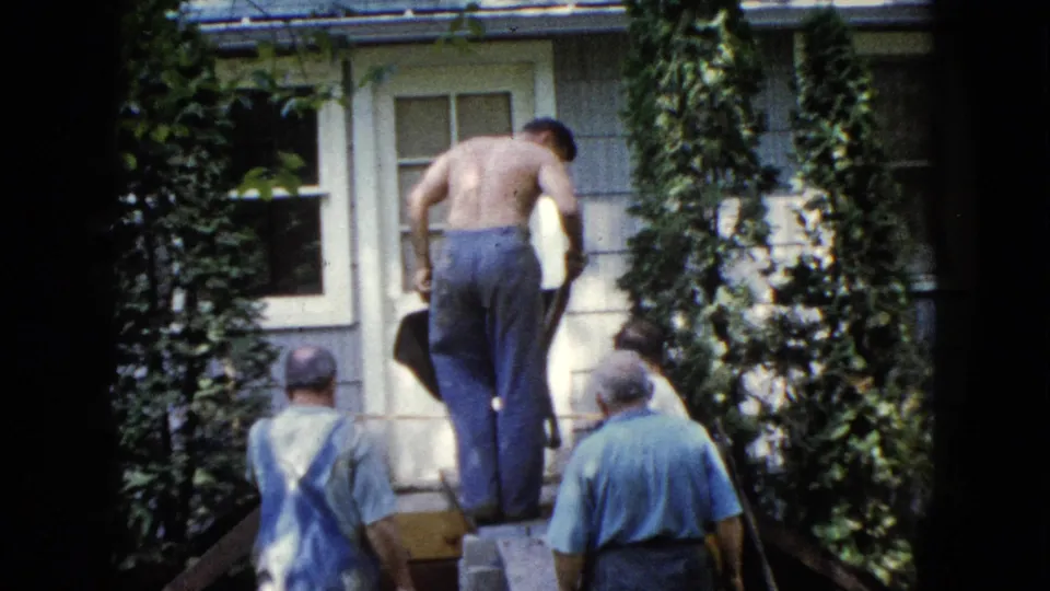 A 1960 Wauconda frame shows a shirtless man on planks at a house entrance while men below observe the outdoor work.