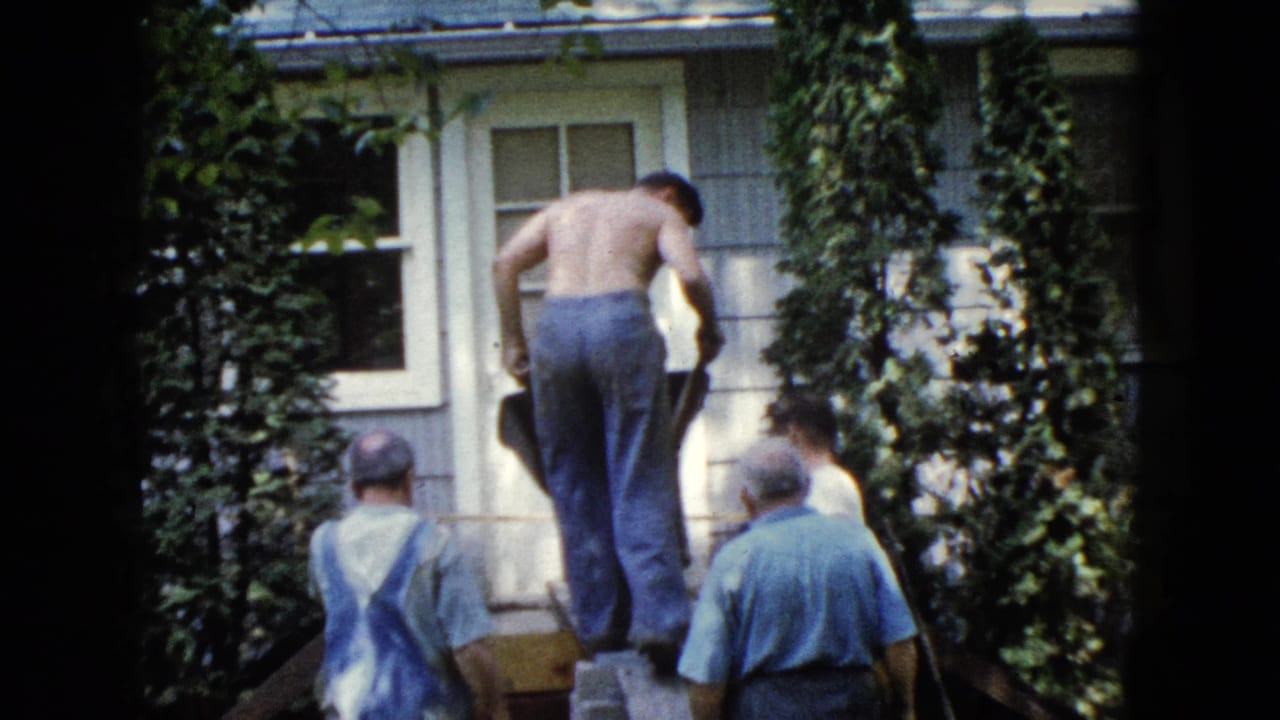 Men gather by a house as a shirtless man moves a wheelbarrow up a wooden ramp.