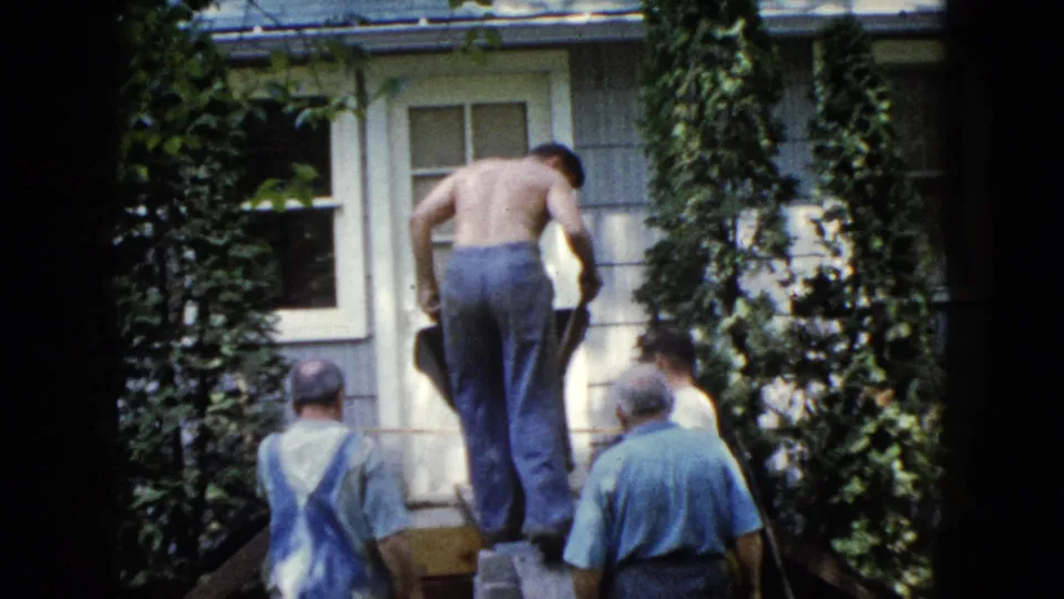 1960 Wauconda home-movie frame showing men outside a house while a shirtless man guides a wheelbarrow up a ramp.