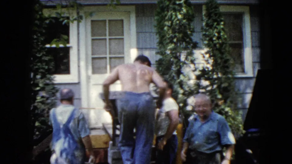 1960 Wauconda frame with men clustered around a ramp and wheelbarrow outside an ivy-covered house exterior.