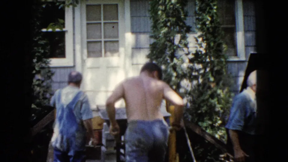 1960 Wauconda home-movie frame showing men repositioning around a wooden ramp beside a light-colored residential exterior.