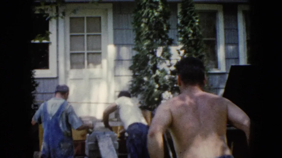1960 Wauconda frame showing men continuing ramp work outside the house, with the shirtless man in the foreground.