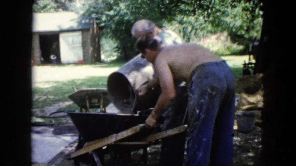 A shirtless worker bends over a wheelbarrow while handling material in a 1960 Wauconda backyard work scene.