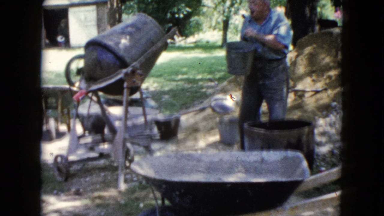 1960 Wauconda home-movie frame showing a man by a concrete mixer in a yard with wheelbarrows, buckets, trees, and an outbuilding.