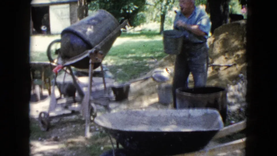 1960 Wauconda home-movie frame showing a man by a concrete mixer in a yard with wheelbarrows, buckets, trees, and an outbuilding.