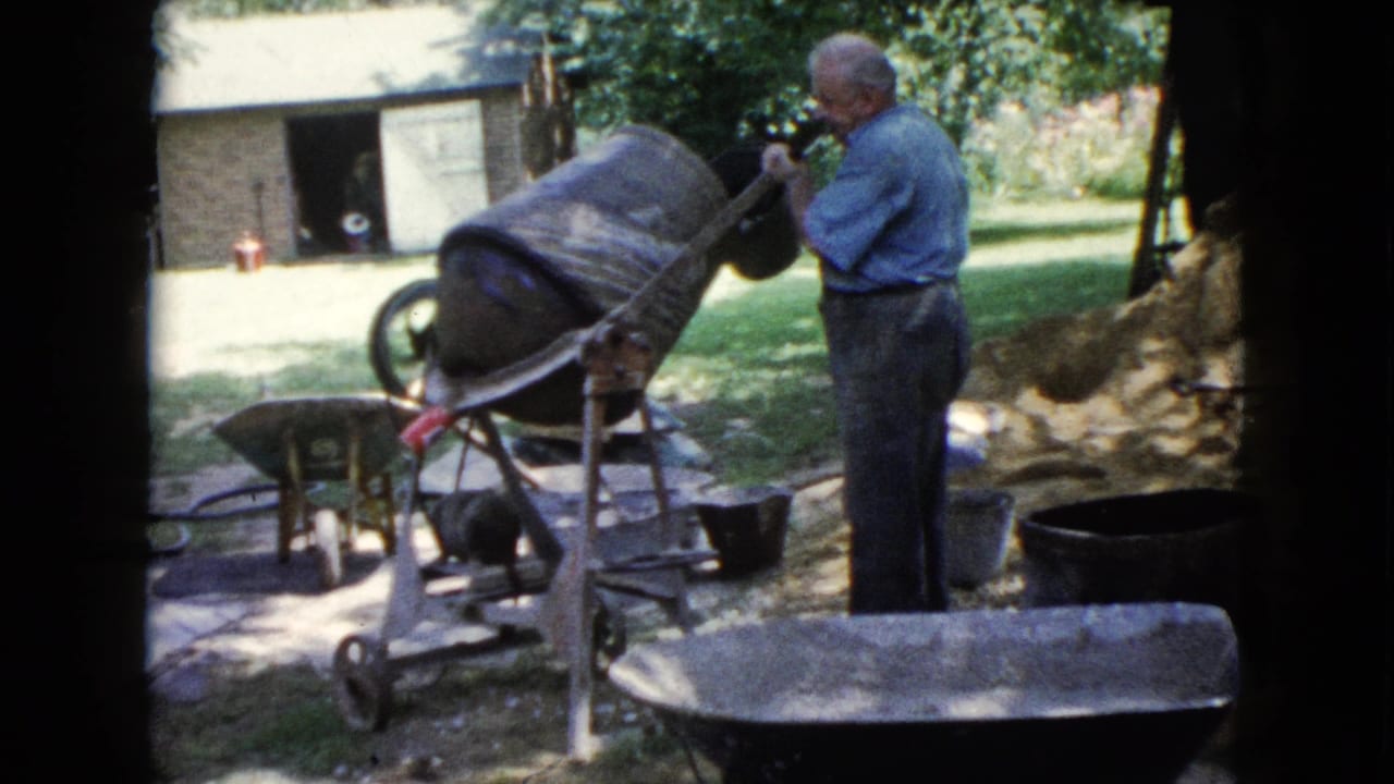 1960 Wauconda frame of an adult man operating a tilted concrete mixer near wheelbarrows, buckets, grass, and a small outbuilding.