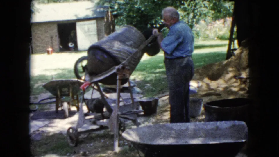 1960 Wauconda frame of an adult man operating a tilted concrete mixer near wheelbarrows, buckets, grass, and a small outbuilding.