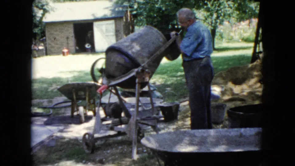 1960 Wauconda home-movie frame showing a man in a blue shirt using a concrete mixer beside wheelbarrows and buckets in a backyard.