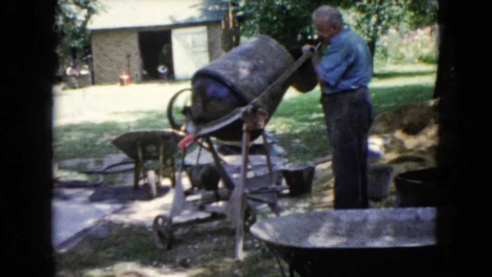 1960 Wauconda frame showing an adult man beside a concrete mixer, with wheelbarrows, buckets, grass, trees, and an outbuilding in view.