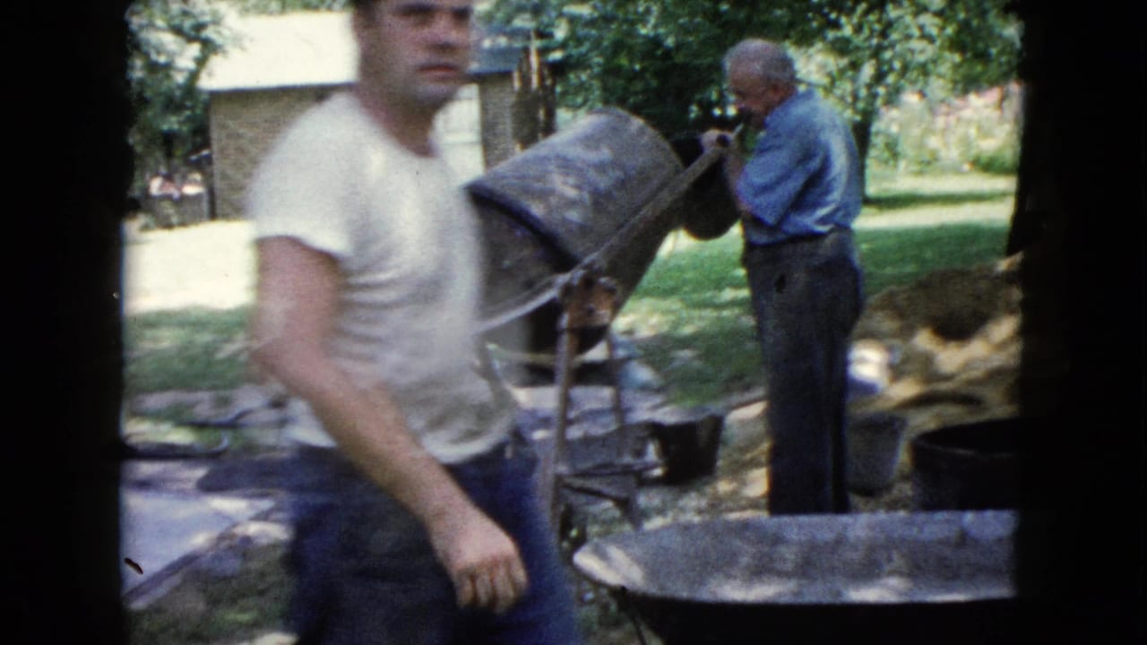 Younger man walks past while older man works beside a concrete mixer in a backyard.