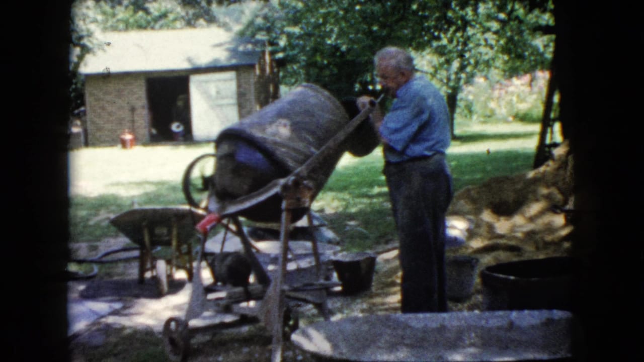 Older man operates a concrete mixer near wheelbarrows and a brick shed.