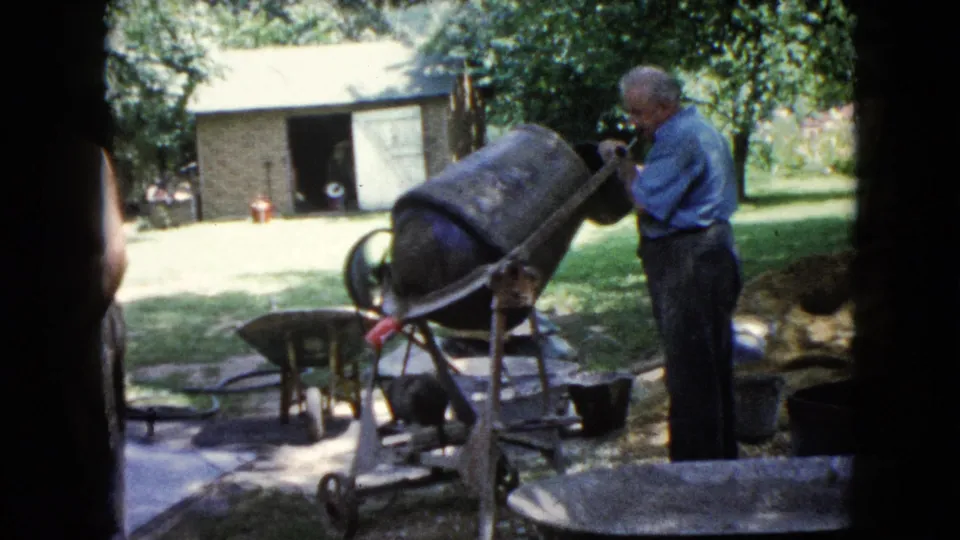 The 1960 home-movie frame shows the older man at the mixer, with a wheelbarrow, brick shed, lawn, and shaded trees around him.