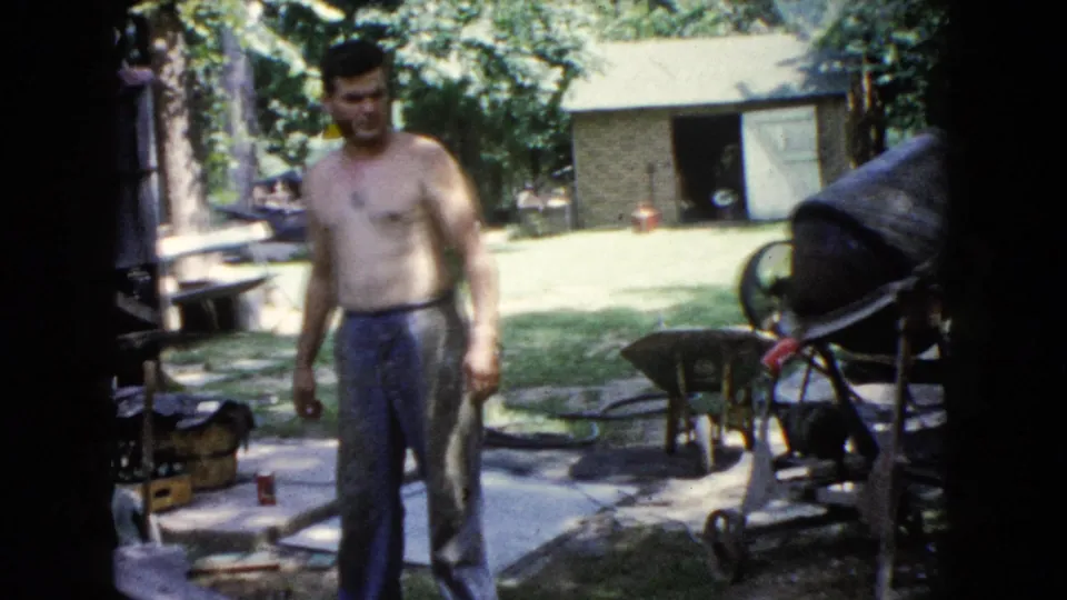 A shirtless younger man faces the camera near the backyard concrete mixer, with a shed, trees, wheelbarrow, and scattered materials visible.