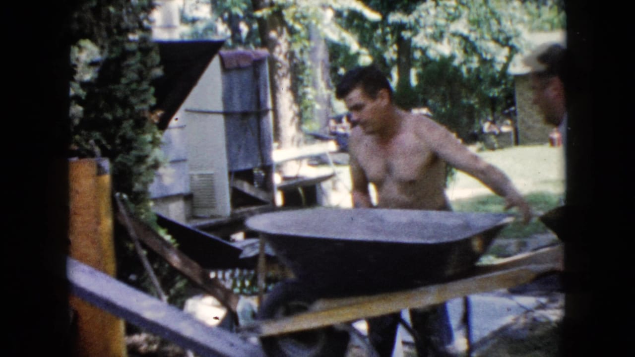 A shirtless man works beside a wheelbarrow and wooden ramp in a Wauconda backyard, with trees and an outbuilding behind him.