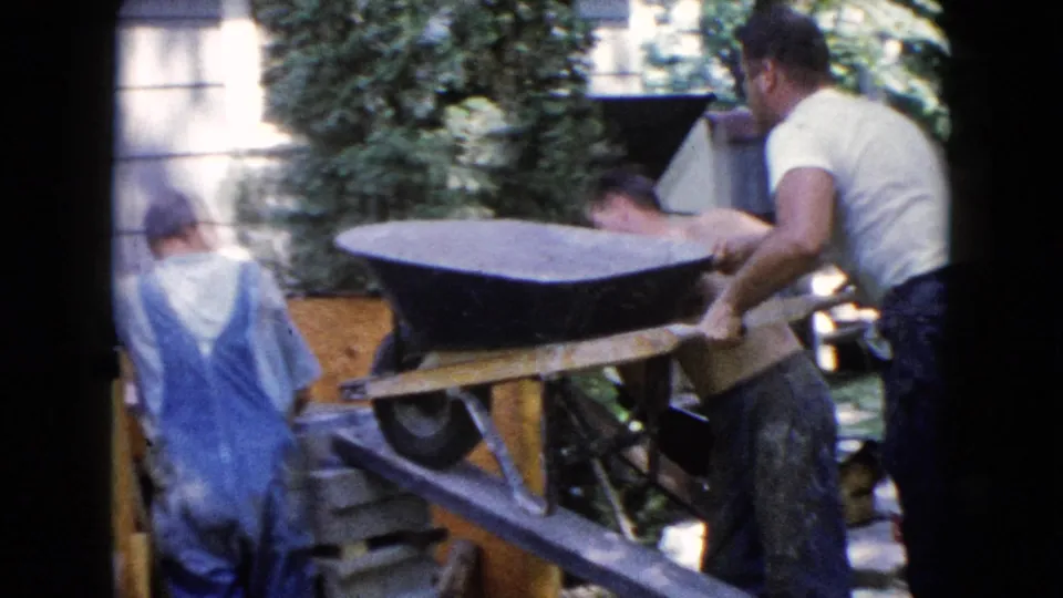 Several adult men in casual work clothes and overalls help move a wheelbarrow up a wooden ramp in the Wauconda backyard scene.