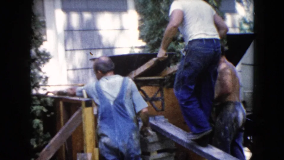 Adult men continue guiding the wheelbarrow up the ramp beside an outbuilding in 1960 Wauconda home-movie footage.