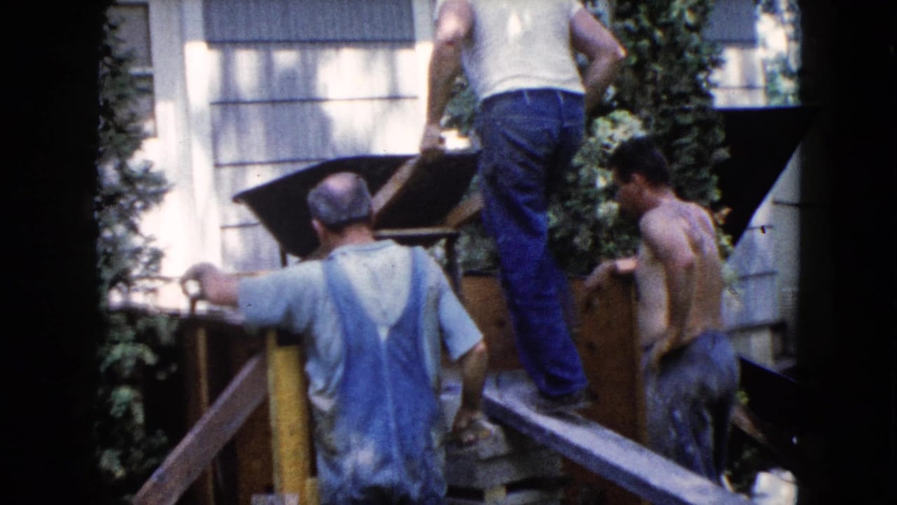 Three men work around a wheelbarrow and wooden platform beside a house.