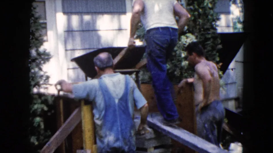 In Wauconda, Illinois, three adult men work beside a house with a wheelbarrow and wood planks in 1960 home-movie footage.
