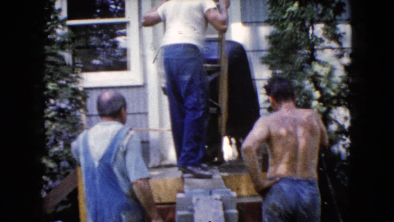 A man stands on a platform while two others watch near a wheelbarrow.